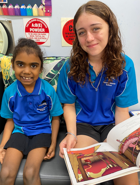 An older Dundee Beach School student reading a book with a younger student